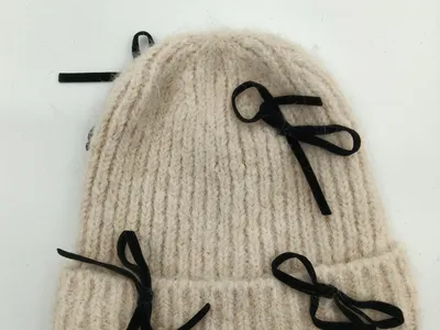 Dicke cremeweiße Wintermütze aus grobem Rippenstrick mit mehreren dekorativen schwarzen Samtbandschleifen, flach auf weißem Hintergrund fotografiert