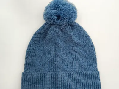 Stahlblaue Zopfstrickmütze mit fluffigem Bommel auf der Spitze und breitem Rippbündchen in Draufsicht auf hellem Hintergrund