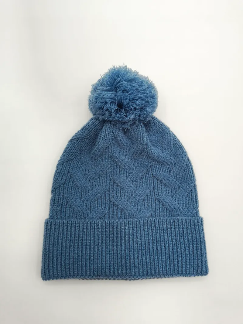 Stahlblaue Zopfstrickmütze mit fluffigem Bommel auf der Spitze und breitem Rippbündchen in Draufsicht auf hellem Hintergrund
