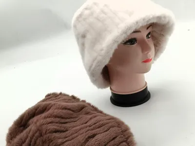 Weißer Kunstfell-Bucket-Hat auf Schaufensterkopf neben flach liegender brauner Plüschmütze auf weißer Fläche
