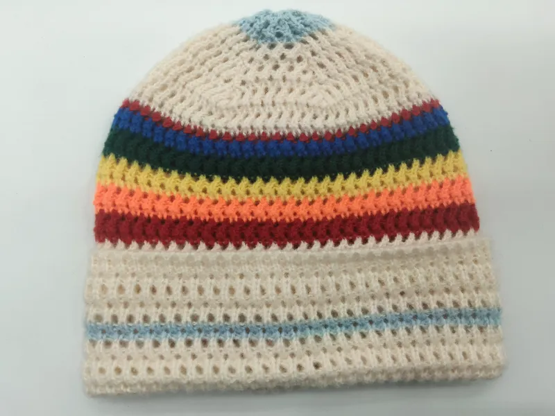 Handgehäkelte Mütze mit bunten Regenbogenstreifen auf cremefarbenem Lochmuster-Grund, auf weißer Fläche liegend