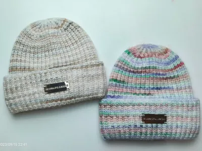 Zwei Grobstrickmützen mit umgeschlagenem Bündchen: eine in neutralem Melange-Beige, eine in pastell-buntem Streifenmuster, beide mit Metalletiketten