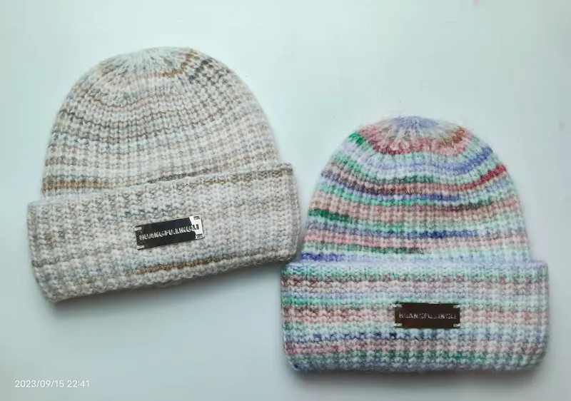 Zwei Grobstrickmützen mit umgeschlagenem Bündchen: eine in neutralem Melange-Beige, eine in pastell-buntem Streifenmuster, beide mit Metalletiketten