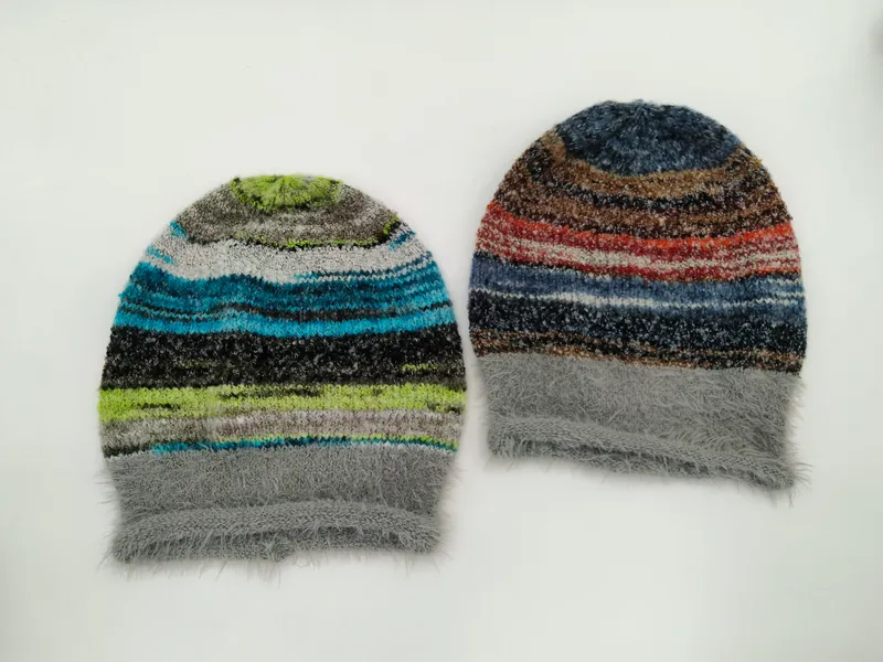 Zwei flauschige Strickmützen mit buntem Streifenmuster, eine in Blau-Grün-Tönen und eine in Rot-Braun-Tönen, beide mit grauem Kunstfellbündchen