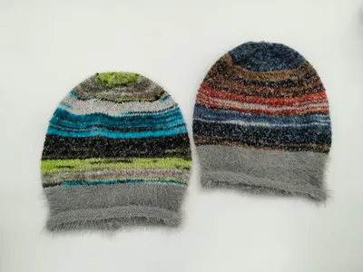 Zwei flauschige Strickmützen mit buntem Streifenmuster, eine in Blau-Grün-Tönen und eine in Rot-Braun-Tönen, beide mit grauem Kunstfellbündchen