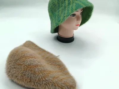 Grün gestreifter flauschiger Strick-Sonnenhut auf einer Hutform zusammen mit einem beigefarbenen flachen Strickhut auf weißem Untergrund