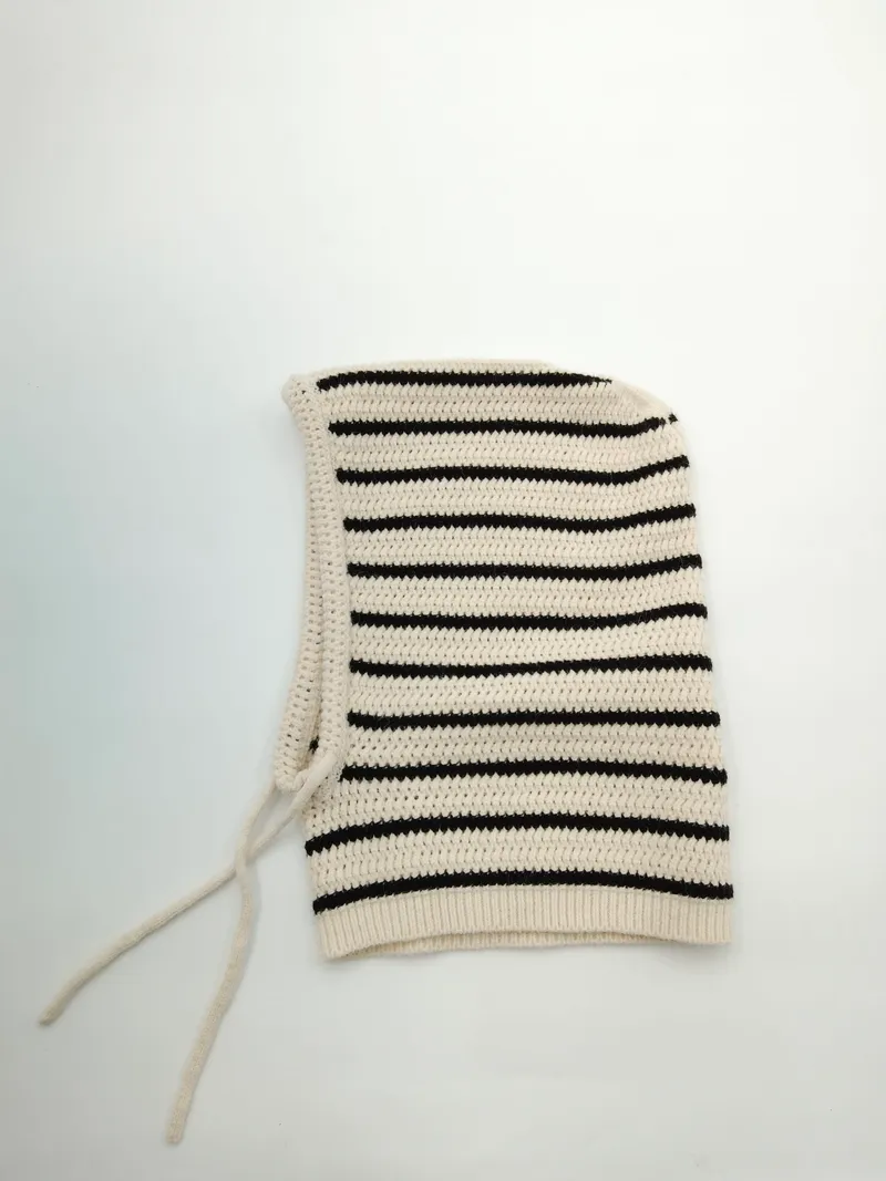 Handgestrickte Sturmhaube aus Baumwolle in Creme und Schwarz mit waagerechten Streifen und verstellbaren Kinnbändern, Draufsicht auf hellem Untergrund