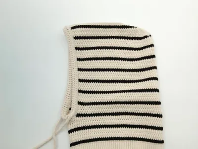 Handgestrickte Sturmhaube aus Baumwolle in Creme und Schwarz mit waagerechten Streifen und verstellbaren Kinnbändern, Draufsicht auf hellem Untergrund