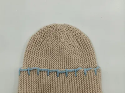 Cremefarbene Wintermütze aus dickem Garn mit auffälliger blauer Kontrastnaht entlang des gefalteten Randes, Studioaufnahme