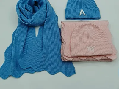 Blauer Strickschal und Mütze mit Buchstaben-A-Stickerei neben rosa Mützen- und Schal-Set mit Schmetterlings-Motiv auf weißem Untergrund