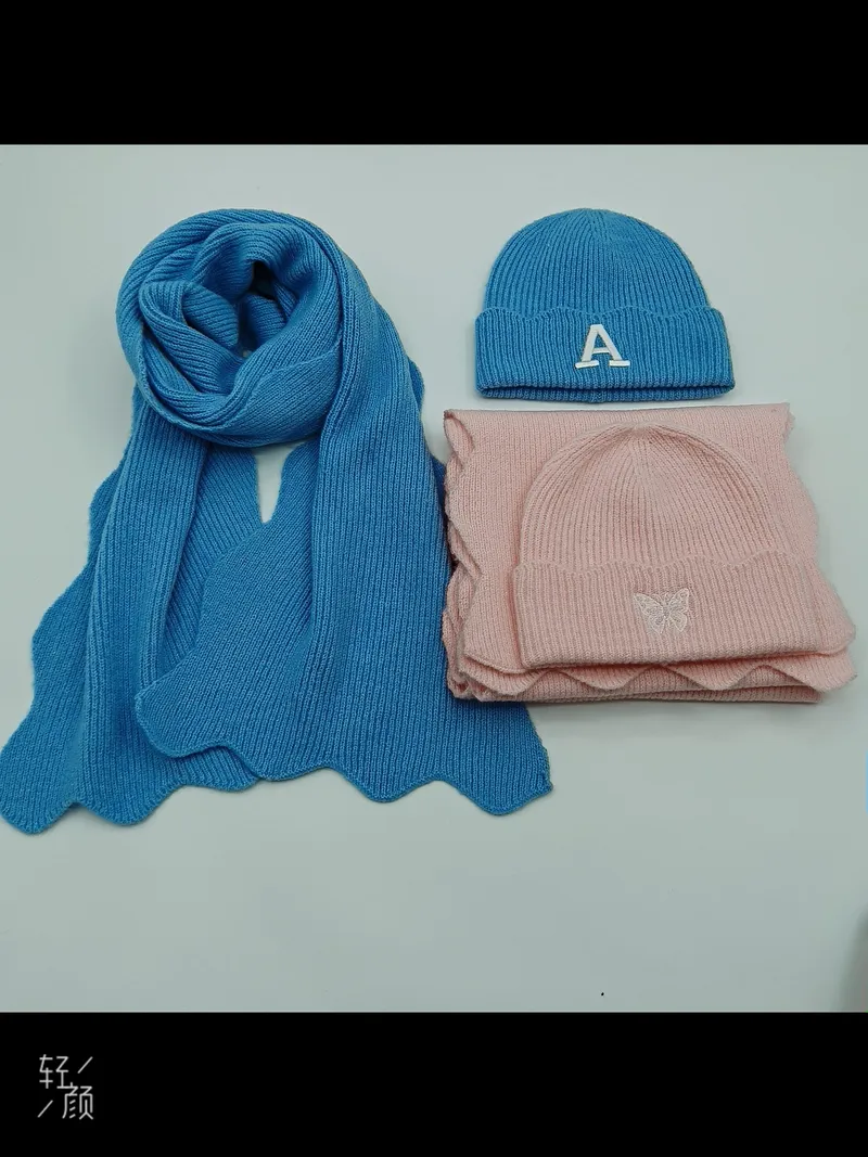 Blauer Strickschal und Mütze mit Buchstaben-A-Stickerei neben rosa Mützen- und Schal-Set mit Schmetterlings-Motiv auf weißem Untergrund