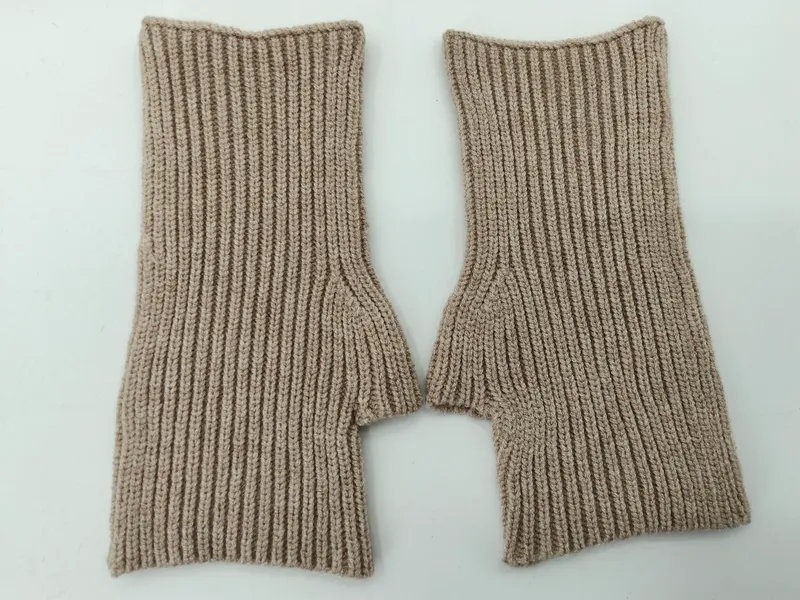 Ein Paar fingerlose Strickhandschuhe in Sandbeige mit Rippenstruktur und Daumenöffnung auf weißem Hintergrund