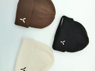 Drei gefaltete Umschlag-Beanies in Schokobraun, Schwarz und Naturweiß mit kleinem eingesticktem Logo auf hellem Untergrund