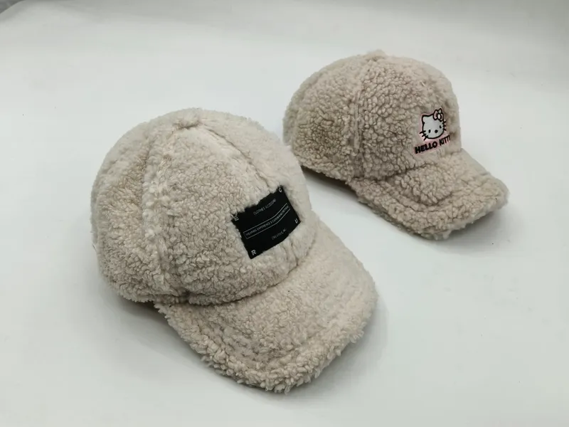 Zwei cremefarbene Teddy-Fleece-Schirmmützen nebeneinander, eine mit schwarzem Label-Aufnäher, die andere mit Hello-Kitty-Stickerei