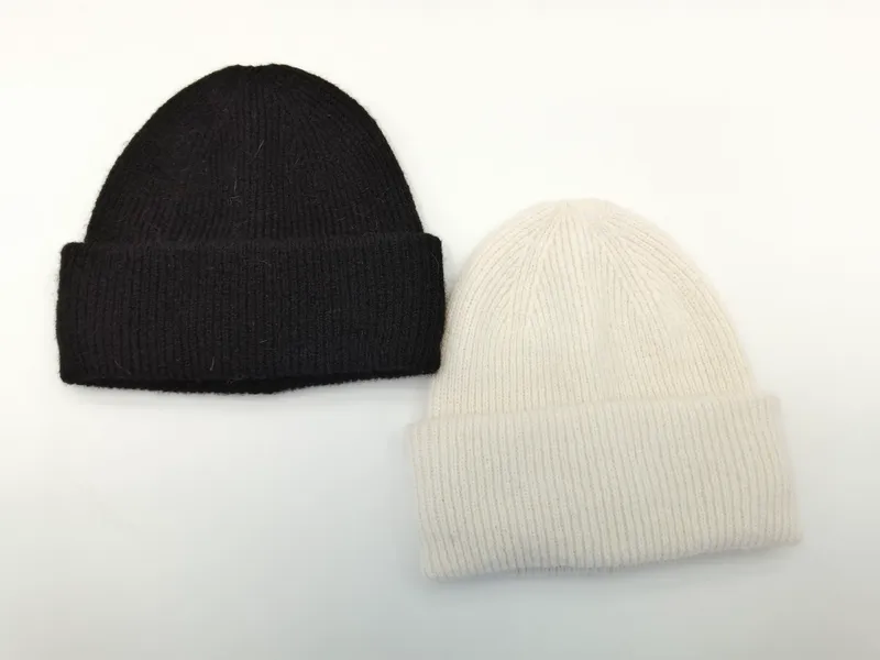 Zwei klassische Rippstrickmützen mit Umschlagrand nebeneinander – eine in Tiefschwarz, eine in gebrochenem Weiß, auf hellem Untergrund