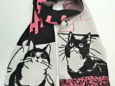 Gestrickter Schal mit schwarz-weißen Smoking-Katzen-Illustrationen und pinkfarbenen geometrischen Akzenten, aufgerollt auf hellem Hintergrund