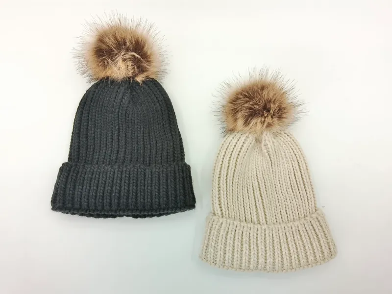 Zwei voluminöse Strickmützen mit großer Kunstfellbommel in Dunkelgrau und hellem Beige auf weißem Hintergrund