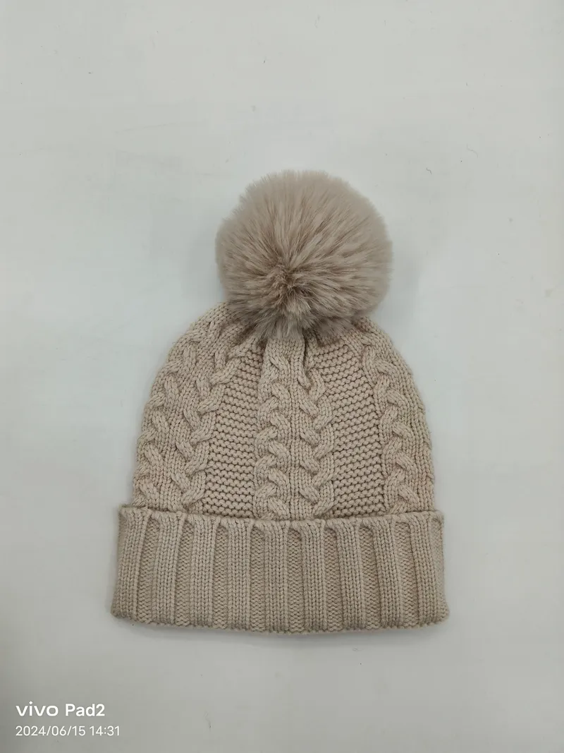 Beige Wintermütze mit klassischem Zopfstrickmuster, großer Kunstfellbommel und geripptem Umschlagrand auf weißem Untergrund