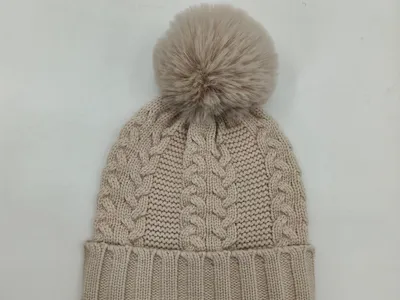 Beige Wintermütze mit klassischem Zopfstrickmuster, großer Kunstfellbommel und geripptem Umschlagrand auf weißem Untergrund