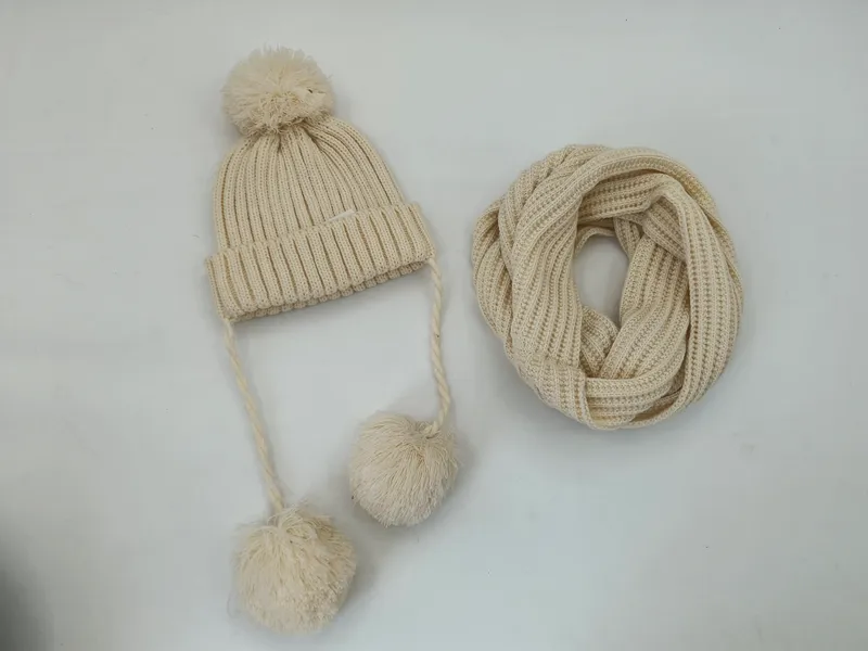 Cremefarbene Rippenstrickmütze mit Bommeln und Zopfbändern sowie passender Loop-Schal, Gesamtansicht auf weißem Hintergrund