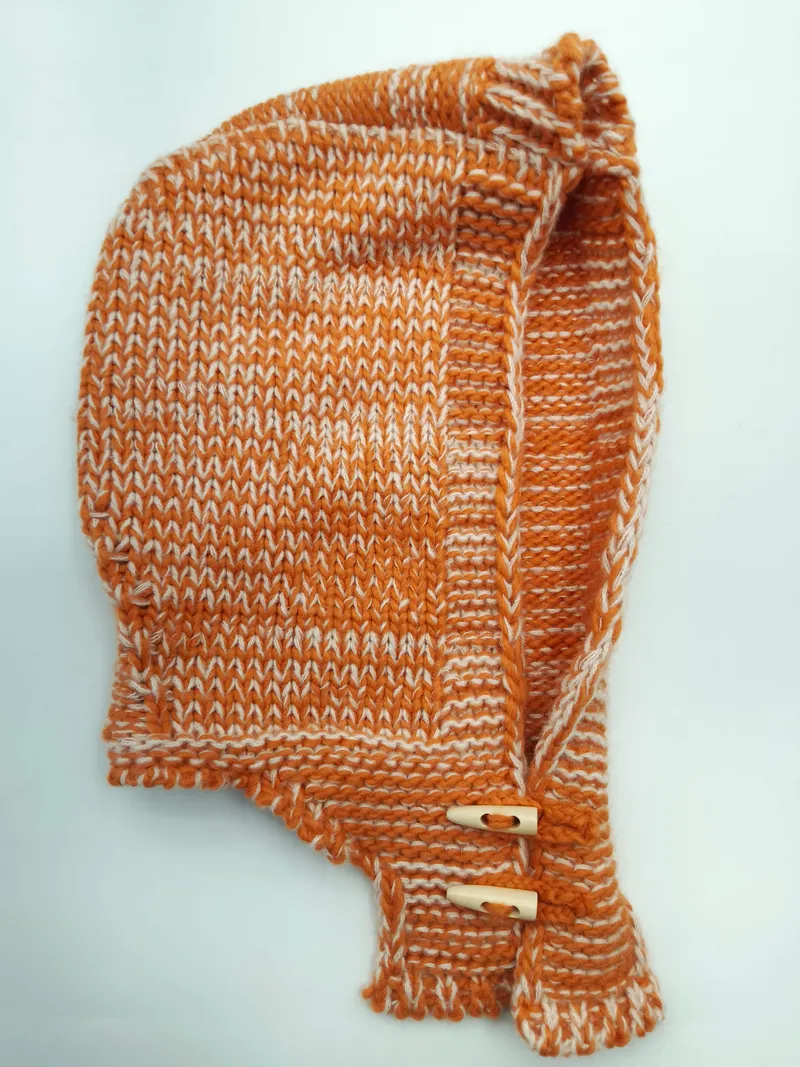 Handgestrickte Säuglingsmütze in rostoranger Melange-Wolle mit cremefarbenen Akzenten und Holzknöpfen an den Bindebändern