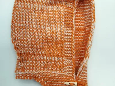 Handgestrickte Säuglingsmütze in rostoranger Melange-Wolle mit cremefarbenen Akzenten und Holzknöpfen an den Bindebändern