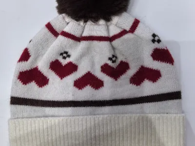 Weiße Strickmütze mit roten Herzchen-Muster, dunkelbraunem Kunstfell-Pompon und geripptem Umschlagrand auf weißem Hintergrund