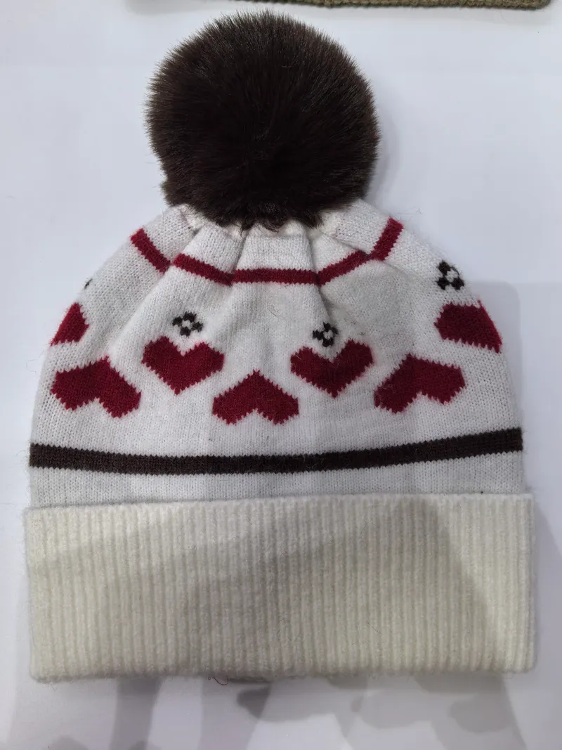 Weiße Strickmütze mit roten Herzchen-Muster, dunkelbraunem Kunstfell-Pompon und geripptem Umschlagrand auf weißem Hintergrund