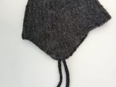 Handgestrickte Babymütze aus anthrazitgrauer Filzwolle mit Ohrenklappen und geflochtenen Bindeschnüren, auf weißem Hintergrund