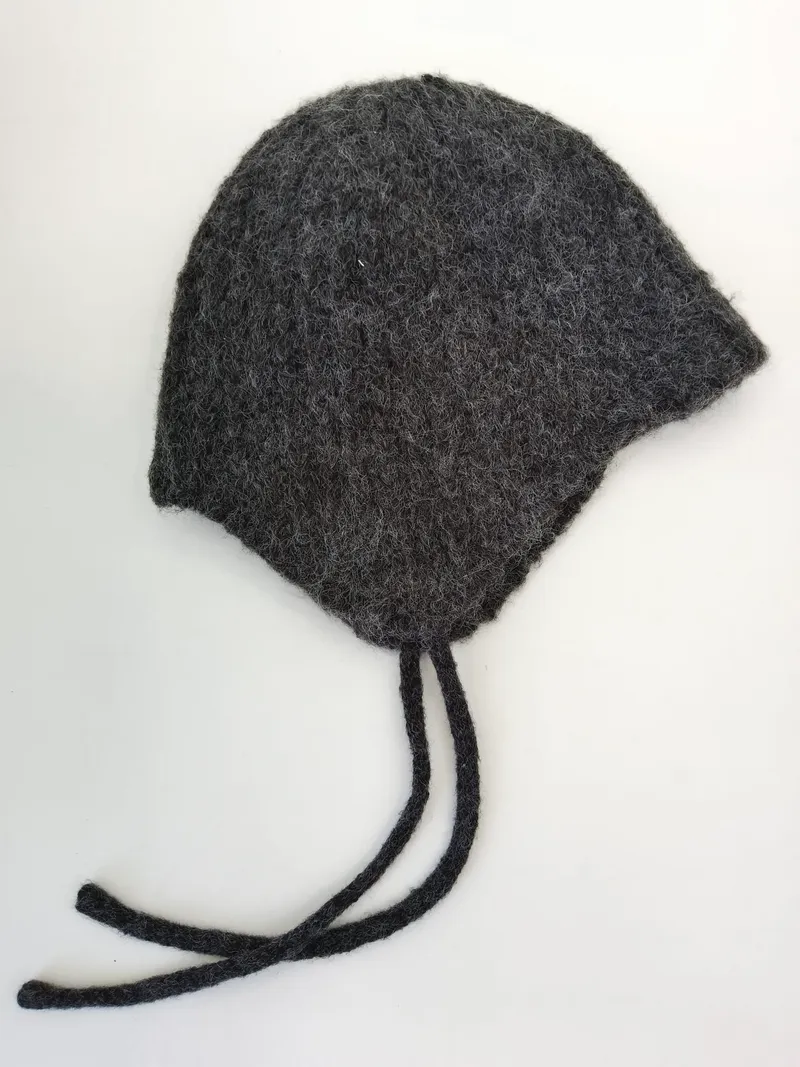 Handgestrickte Babymütze aus anthrazitgrauer Filzwolle mit Ohrenklappen und geflochtenen Bindeschnüren, auf weißem Hintergrund