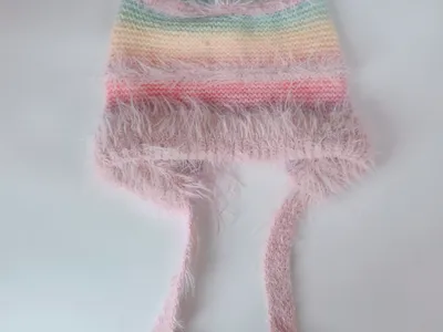 Handgestrickte Baby-Beanie mit pastellfarbenen Streifen, fluffigem rosa Eyelash-Garn-Rand und langen Bindeschnüren, auf hellem Hintergrund