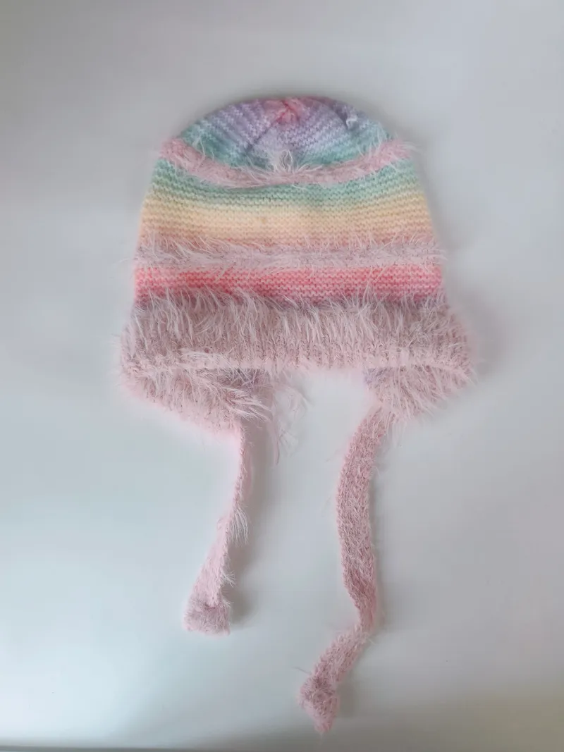 Handgestrickte Baby-Beanie mit pastellfarbenen Streifen, fluffigem rosa Eyelash-Garn-Rand und langen Bindeschnüren, auf hellem Hintergrund