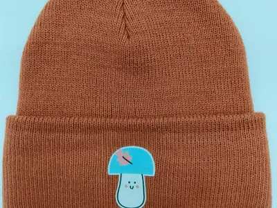 Rostbraune Rippenstrick-Umschlagmütze mit kawaii Pilzfigur als Aufnäher auf der Krempe, flach fotografiert