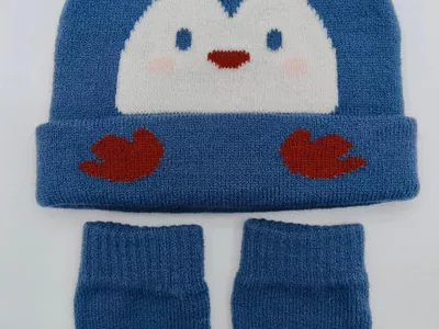 Blaue gestrickte Babymütze mit Intarsia-Pinguin-Gesicht und dazu passenden Babyhandschuhen auf weißem Hintergrund