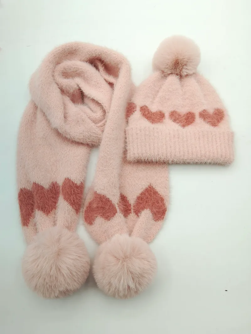 Pinkes flauschiges Winter-Strickset aus Mütze und Schal mit Rosen-Herz-Jacquardmuster und Kunstfell-Bommel-Verzierung auf weißem Hintergrund