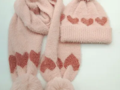 Pinkes flauschiges Winter-Strickset aus Mütze und Schal mit Rosen-Herz-Jacquardmuster und Kunstfell-Bommel-Verzierung auf weißem Hintergrund