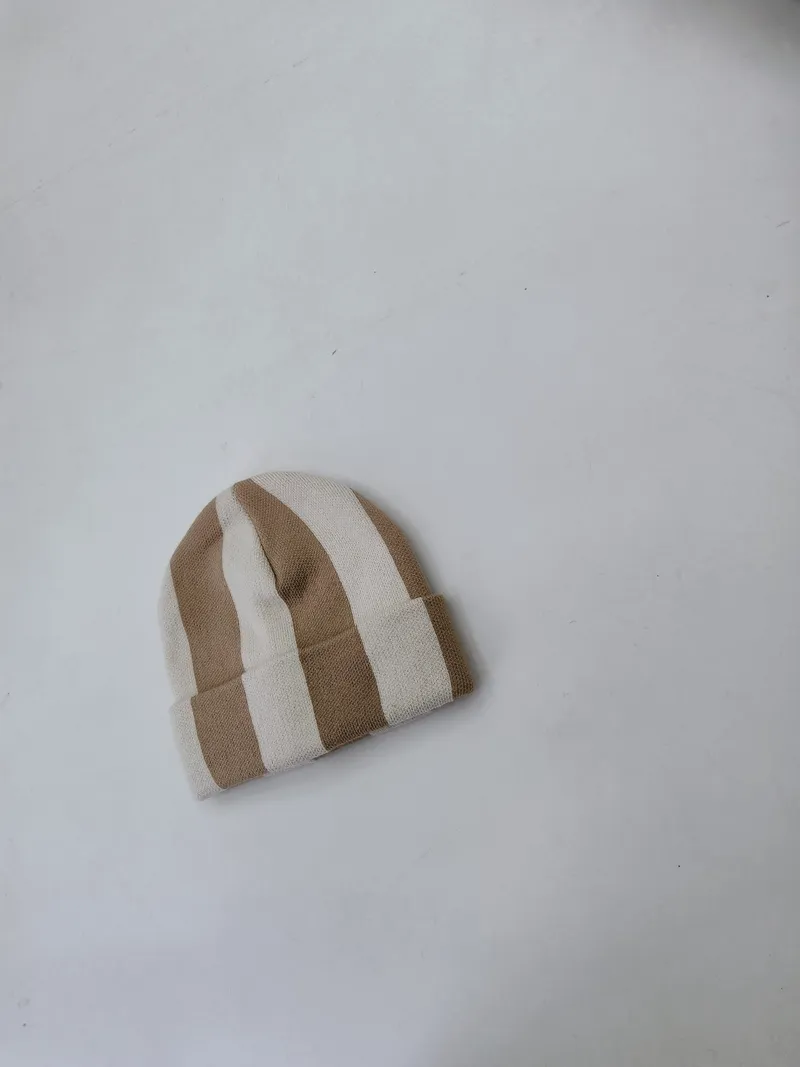 Kleine gestrickte Mütze mit breiten vertikalen Streifen in Beige und Creme, flach auf hellem Untergrund fotografiert