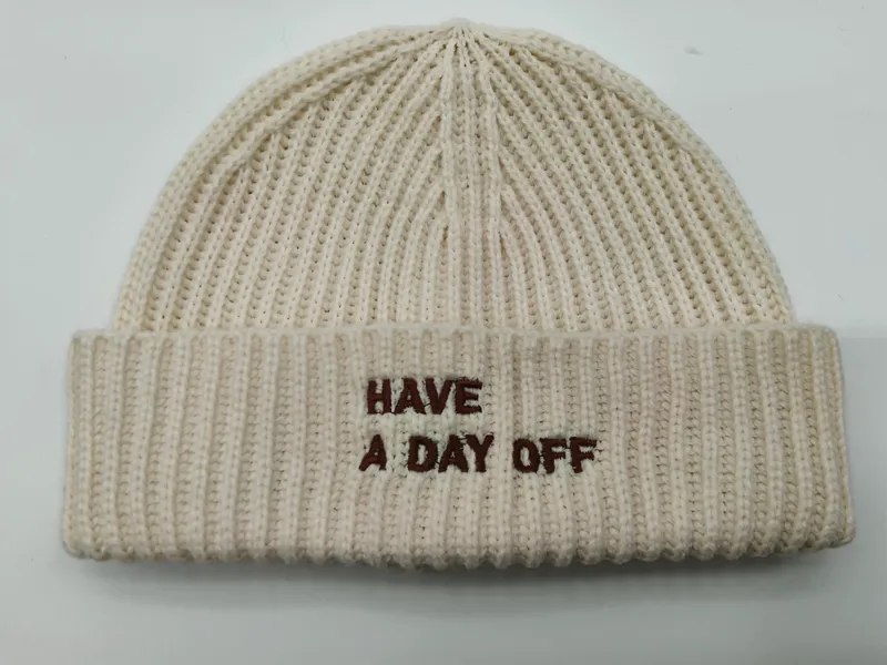 Cremefarbene grob gerippte Strickmütze mit braunem besticktem Schriftzug 'Have A Day Off' auf umgeschlagenem Bund