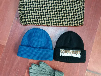 Draufsicht auf beige-schwarzen Hahnentritt-Fransenschal, blaue Waffle-Knit-Beanie, schwarze bestickte Beanie und graue Zopfstrick-Handschuhe auf Holzboden
