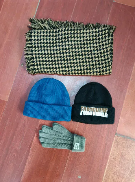 Draufsicht auf beige-schwarzen Hahnentritt-Fransenschal, blaue Waffle-Knit-Beanie, schwarze bestickte Beanie und graue Zopfstrick-Handschuhe auf Holzboden