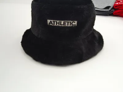 Schwarze flauschige Kunstfell-Fischermütze mit gesticktem Athletic-Etikett-Aufnäher, präsentiert auf weißer Oberfläche mit grauen und roten Accessoires