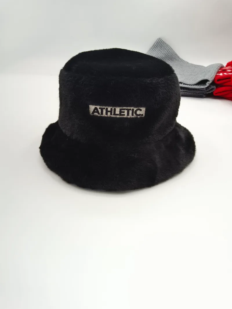 Schwarze flauschige Kunstfell-Fischermütze mit gesticktem Athletic-Etikett-Aufnäher, präsentiert auf weißer Oberfläche mit grauen und roten Accessoires