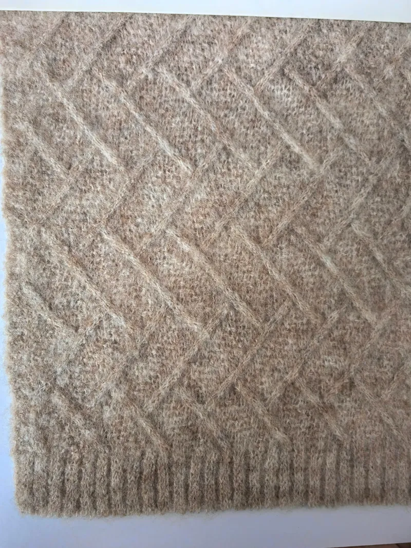 Nahaufnahme einer beigefarbenen Wollmisch-Wurfdecke mit Rautengitterstrickmuster und geripptem Rand