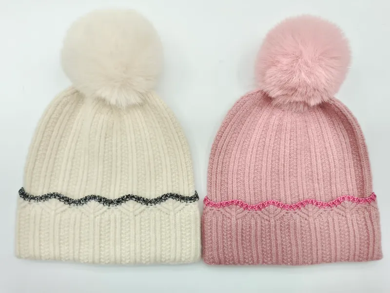 Zwei Strickbommelmützen nebeneinander – eine cremefarben mit schwarzem Glitzerrand und eine rosa mit rosafarbenem Glitzerrand, beide mit Kunstfellbommel