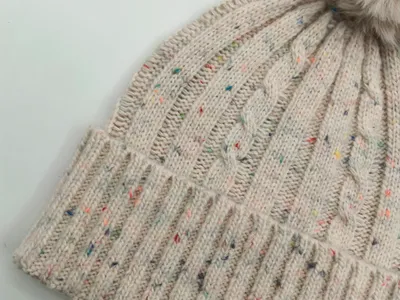 Nahaufnahme einer cremefarbenen Zopfstrickmütze mit bunten Farbsprenkelgarn, Rippenbündchen und flauschigem Kunstfell-Bommel