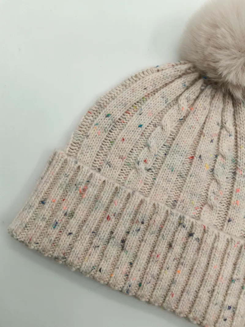 Nahaufnahme einer cremefarbenen Zopfstrickmütze mit bunten Farbsprenkelgarn, Rippenbündchen und flauschigem Kunstfell-Bommel