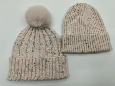 Zwei cremefarbene Strickmützen mit Farbsprenkeln nebeneinander – eine als Zopfstrick mit Kunstfell-Bommel, eine als klassischer Rippenbeanie