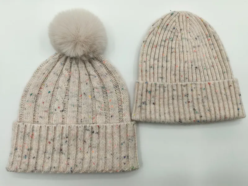 Zwei cremefarbene Strickmützen mit Farbsprenkeln nebeneinander – Zopfstrickvariante mit Kunstfell-Bommel und Rippenstrickvariante mit Umschlagrand