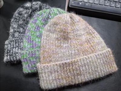 Drei unterschiedliche handgestrickte Wintermützen in Graumeliert, Grün-Lila-Streifen und warmen Beige-Gold-Tönen auf dunklem Untergrund