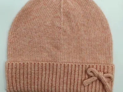 Zarte Strickmütze in Rosa-Melange mit dekorativer Schleife am gefalteten Bündchen, auf hellem Hintergrund